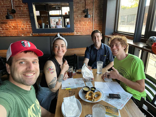 Four people are sitting at a table in a restaurant. Postcards, plates of food, and beverages are on the table in front of them. They are all looking at the camera and smiling.