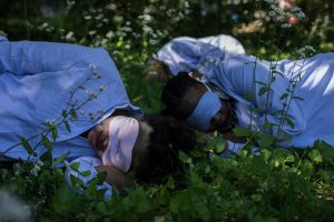 Outfitted in lavender pajamas and sleep masks, people doze off on a bed of ferns in a public demonstration against grind culture. | Photo Credit: The Nap Ministry