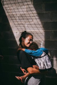 A woman holds a young child close to her chest, gridded sunlight shining down upon them. Photo Credit: Edward Eyer
