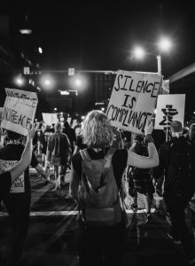 At an urban protest, people walk through the streets with signs exclaiming "No Justice, No Peace" and "Silence is Compliance." Photo Credit: Kelly M Lacy